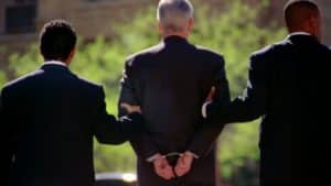 Two detectives in suits escort a man with his hands cuffed behind him.