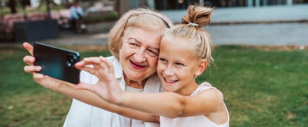 Grandma Taking Selfie With Granddaugter Sitting On Bench. Girl Spending Time With Senior Grandmother, During Summer Brea