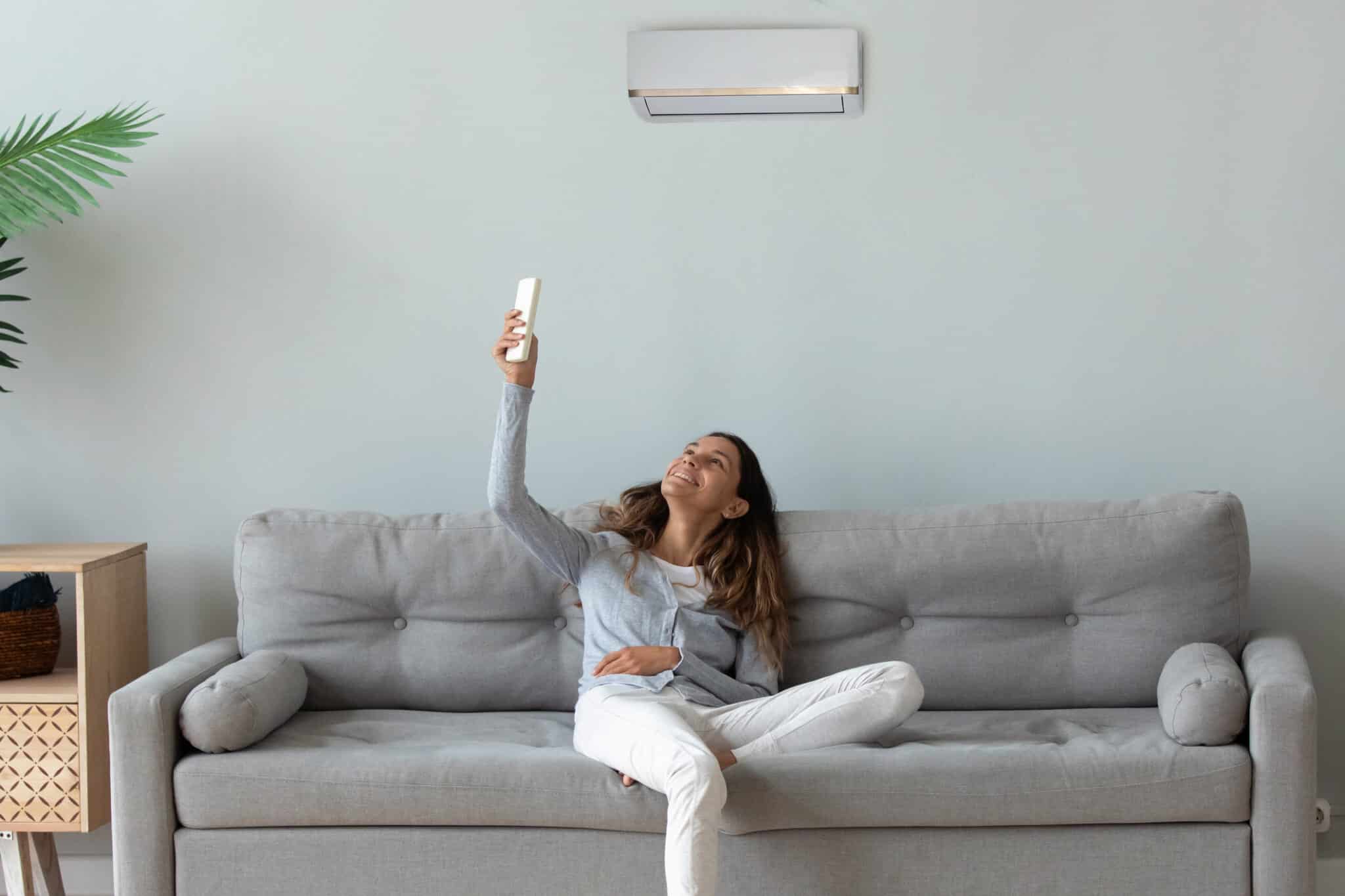 Smiling woman relaxing on couch, using air conditioner remote controller